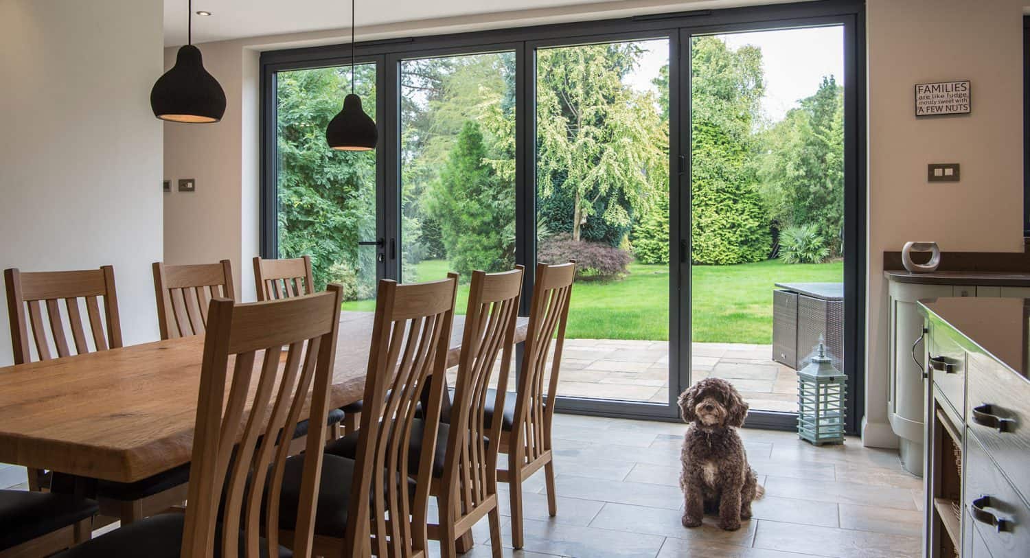 A small brown dog sits on the floor near glass doors in a dining room with a wooden table, chairs, and a view of a green garden outside.