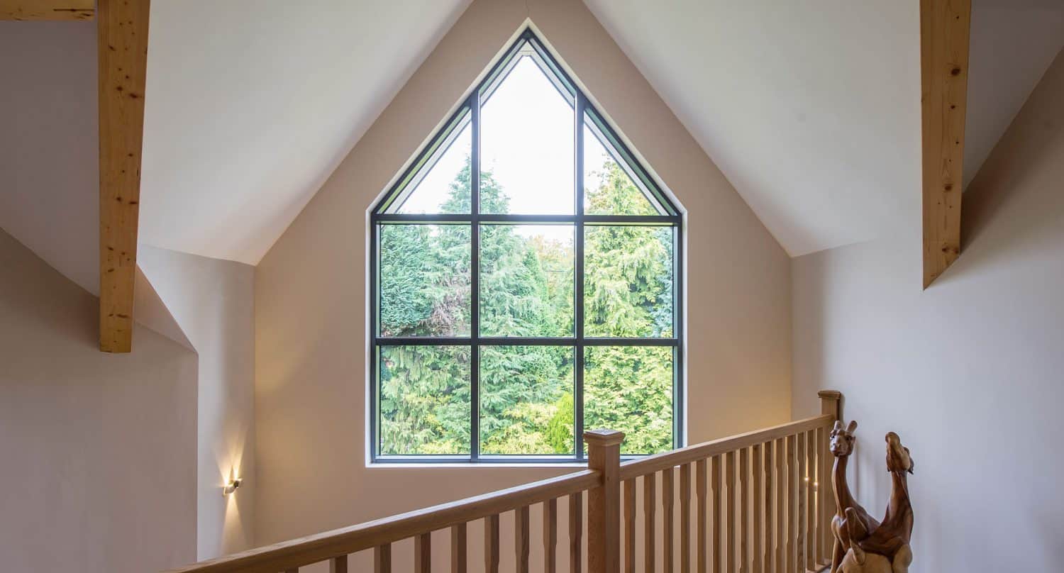 Large triangular window with black frames in a vaulted room, overlooking green trees outside. Wooden railing and beams frame the space, and horse sculptures sit on the landing near the window.