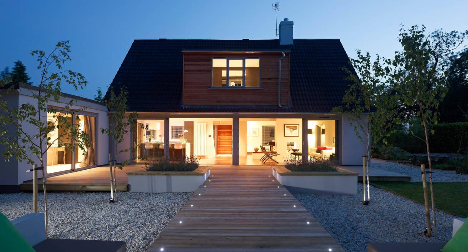 Modern house at dusk with large glass doors, warm indoor lighting, a wooden walkway lined with lights, small trees, and gravel landscaping in the front yard.