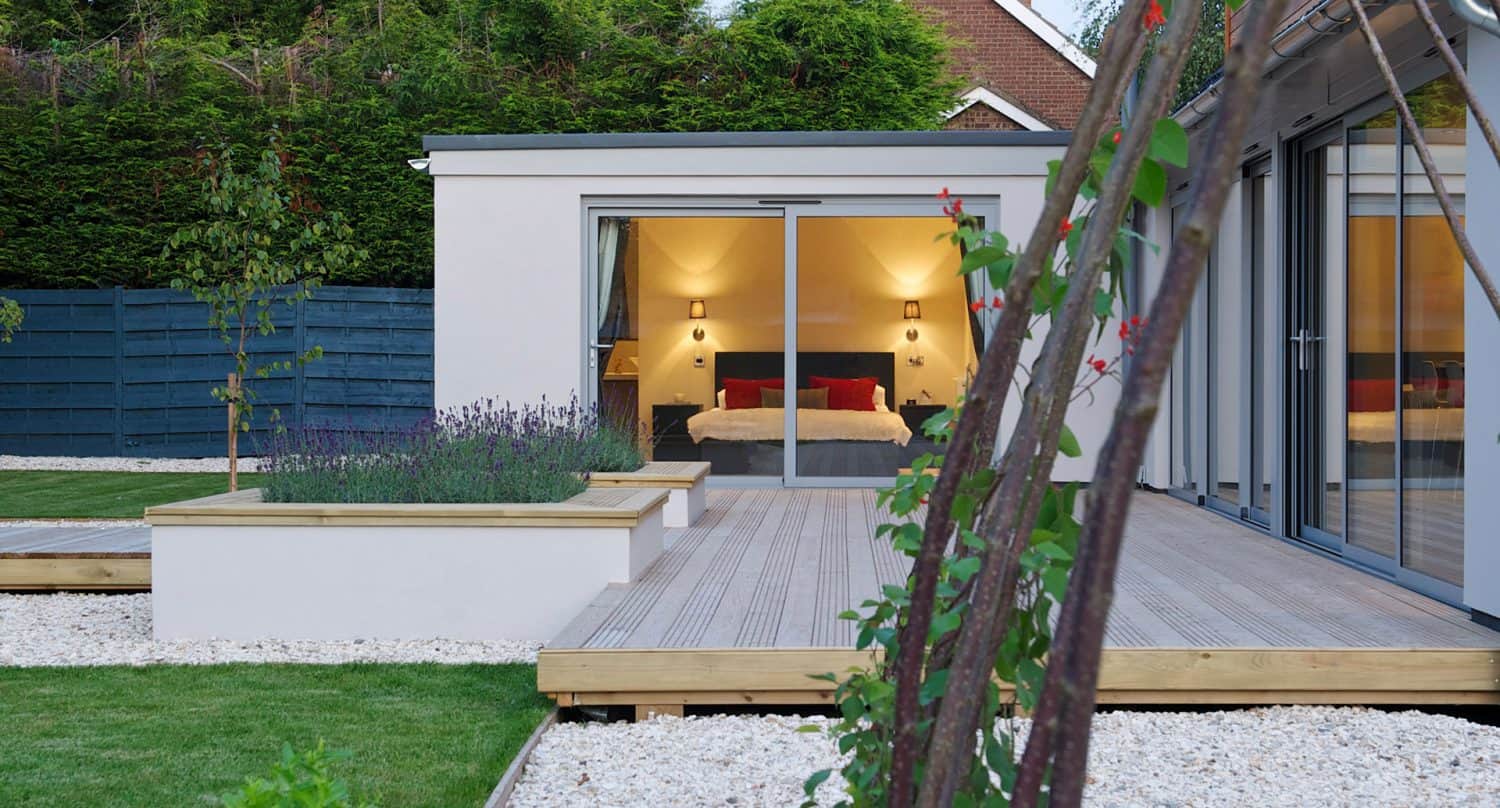 Modern house with large sliding glass doors opening to a deck, revealing a cozy bedroom with warm lighting. The garden features raised beds, gravel, and green grass, with trees and a blue fence in the background.