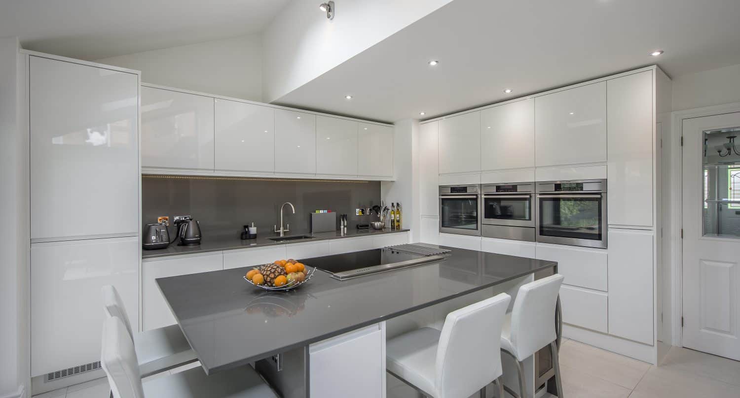 Modern kitchen with white cabinets and grey countertops, featuring an island with four white chairs, built-in ovens, small appliances, and a fruit bowl. Sliding doors fill the bright, spacious area with natural light.