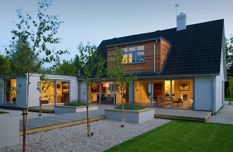 Modern two-story house with large windows, a dark sloped roof, and wood accents. The yard features a wooden deck, gravel landscaping, small trees, and a well-maintained lawn. The home is lit warmly from inside.