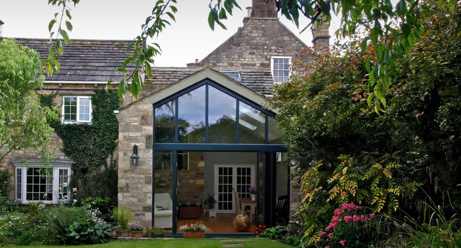 A stone house with a modern glass extension, surrounded by lush greenery, colorful flowers, and a well-kept lawn with stepping stones leading to the entrance.