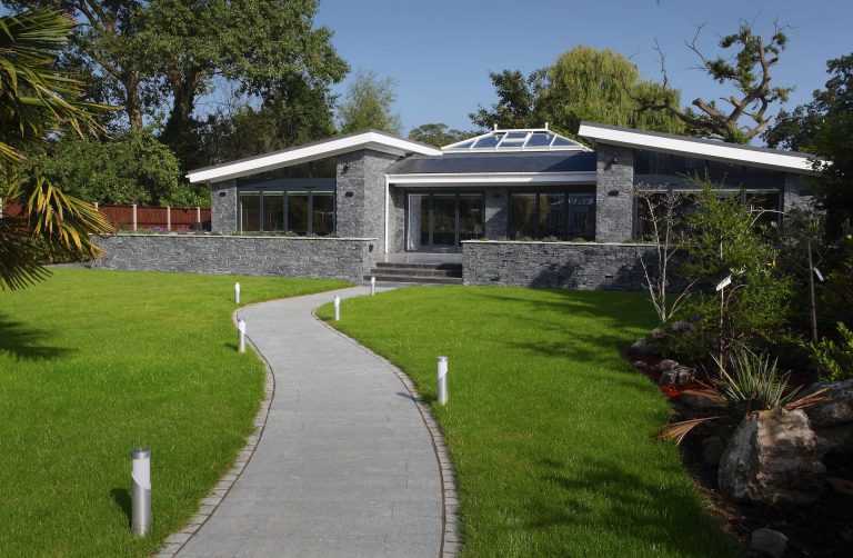Modern single-story house with large windows, dark stone exterior, and a glass roof section. A curved pathway leads through a well-maintained green lawn surrounded by trees and shrubs.