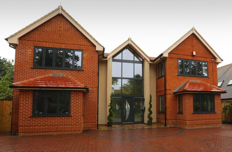 A modern two-story brick house with large dark-framed windows, two front gables, a central glass entrance, and a paved driveway. Two spiral-shaped shrubs stand by the front door.