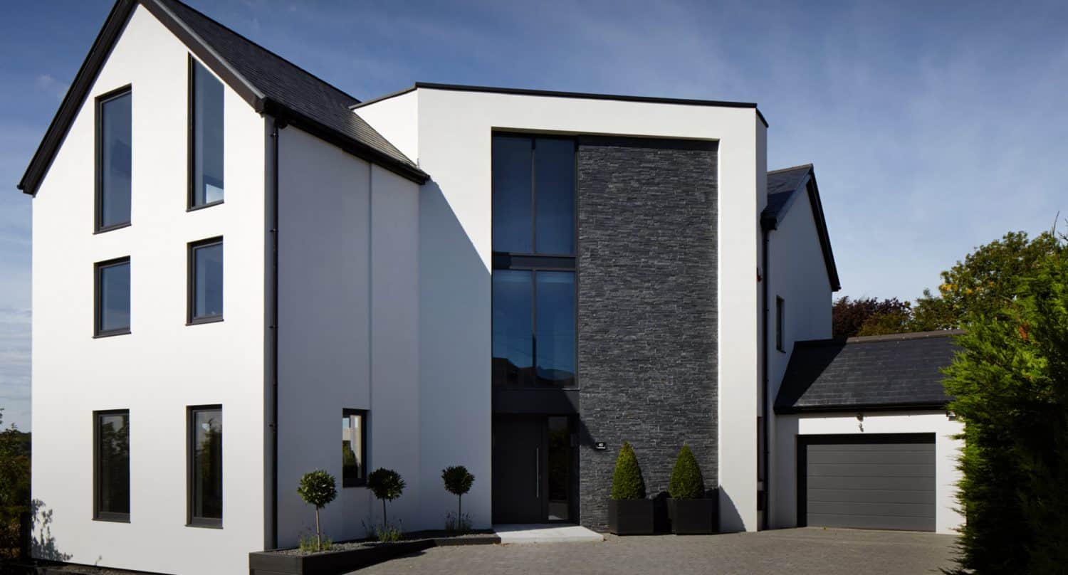Modern two-story house with large windows, white exterior walls, dark stone accents, and a gray garage. The driveway is paved and landscaped with small trees and shrubs. The sky is clear and blue.