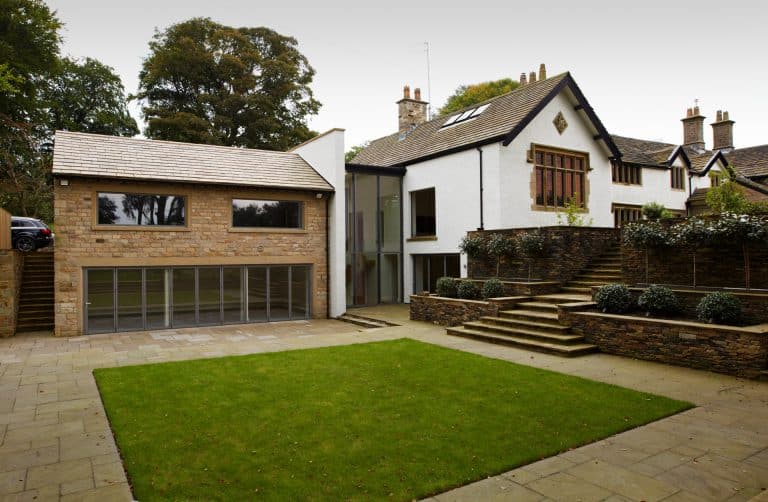 Modern extension with glass doors is attached to a traditional white house with stone accents. There are stone steps, a retaining wall with plants, and a small rectangular lawn in the courtyard.