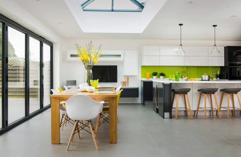 Modern kitchen and dining area with a wooden table, white chairs, large glass doors, a skylight, green backsplash, and a kitchen island with bar stools. Yellow flowers in a vase brighten the table.