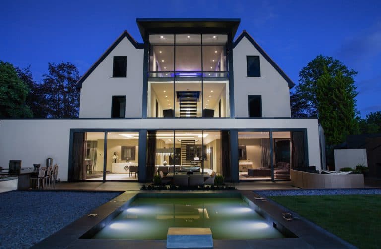 A modern three-story house with large glass windows and a lit interior, viewed at dusk. The house features a central staircase and a rectangular outdoor pool in the foreground, surrounded by outdoor seating and a landscaped yard.