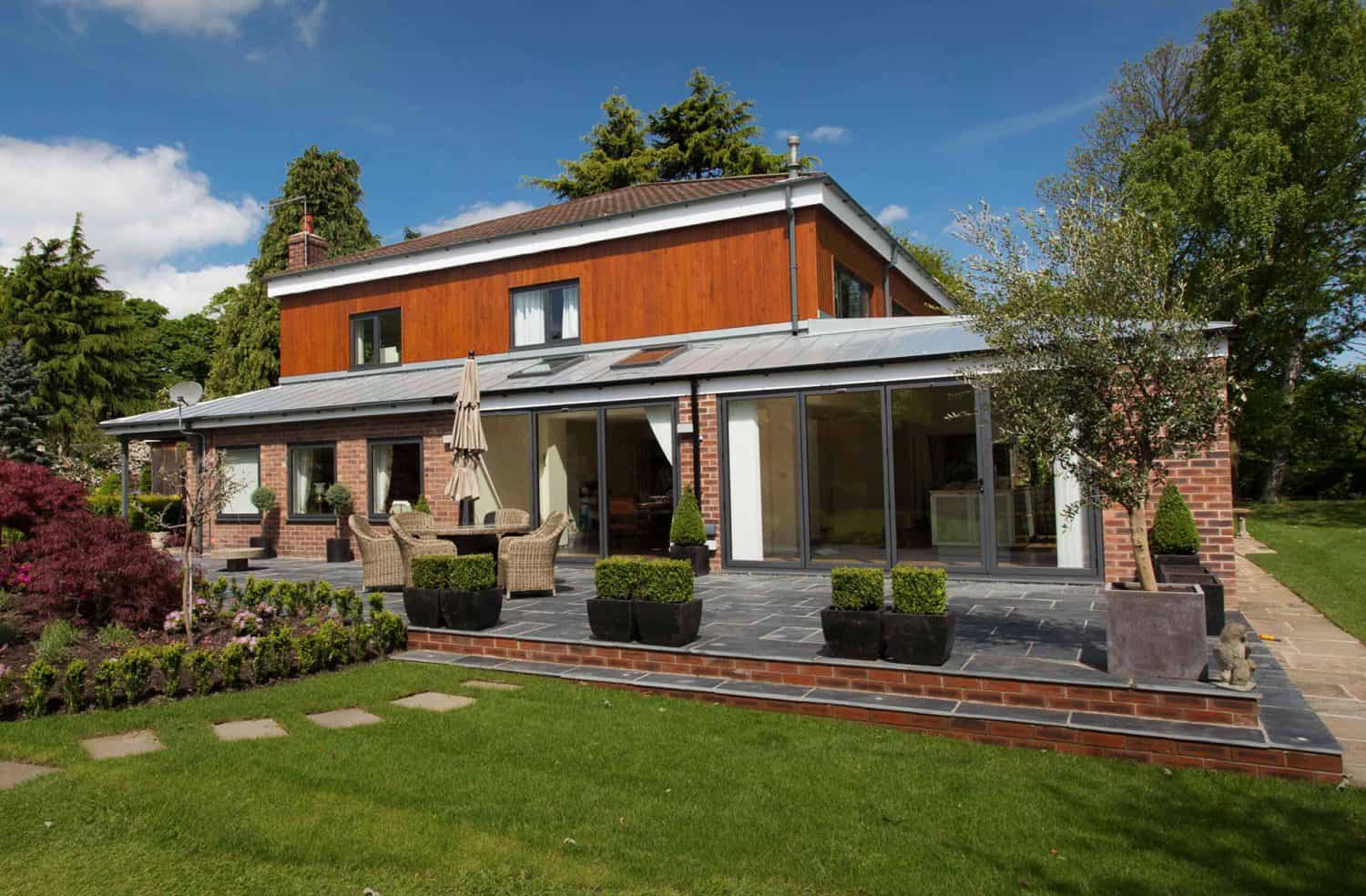 Modern two-story house with brick and wood exterior, large windows, and a patio with outdoor seating. Surrounded by green lawn, potted plants, and trees under a blue sky with clouds.