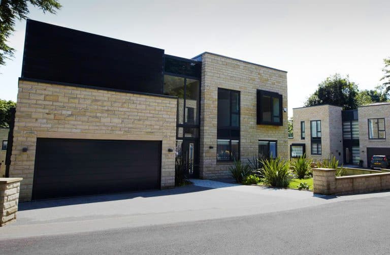 Modern two-story house with light stone exterior, large black-framed windows, and a double garage. The driveway is paved, with landscaped plants in front. Another similar building and a car are visible in the background.