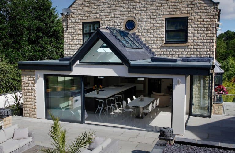 Modern home with a stone exterior and large glass sliding doors opening to a spacious kitchen and dining area. The extension features a sloped glass roof and patio with outdoor seating surrounded by greenery.