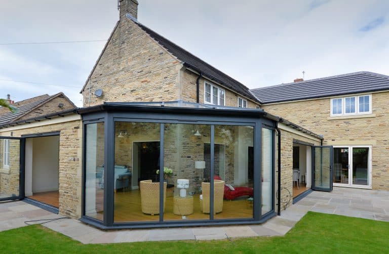 A modern house with stone brick walls features a glass-walled sunroom extension with wicker chairs and a table inside, opening onto a patio and green lawn.