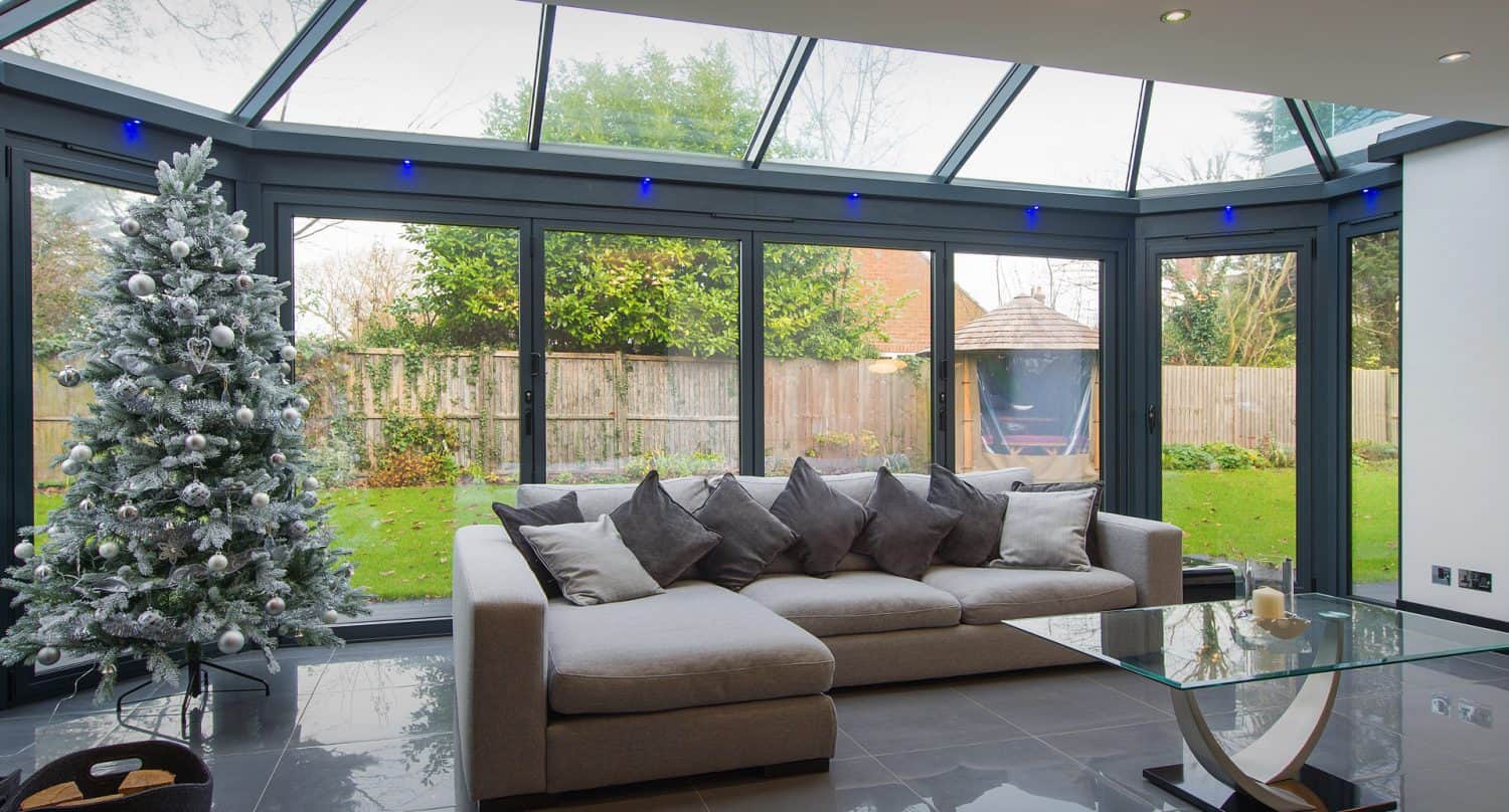A modern sunroom with large glass windows, a light grey sectional sofa with many pillows, a glass coffee table, and a decorated Christmas tree. Outside, a green garden and wooden fence are visible.