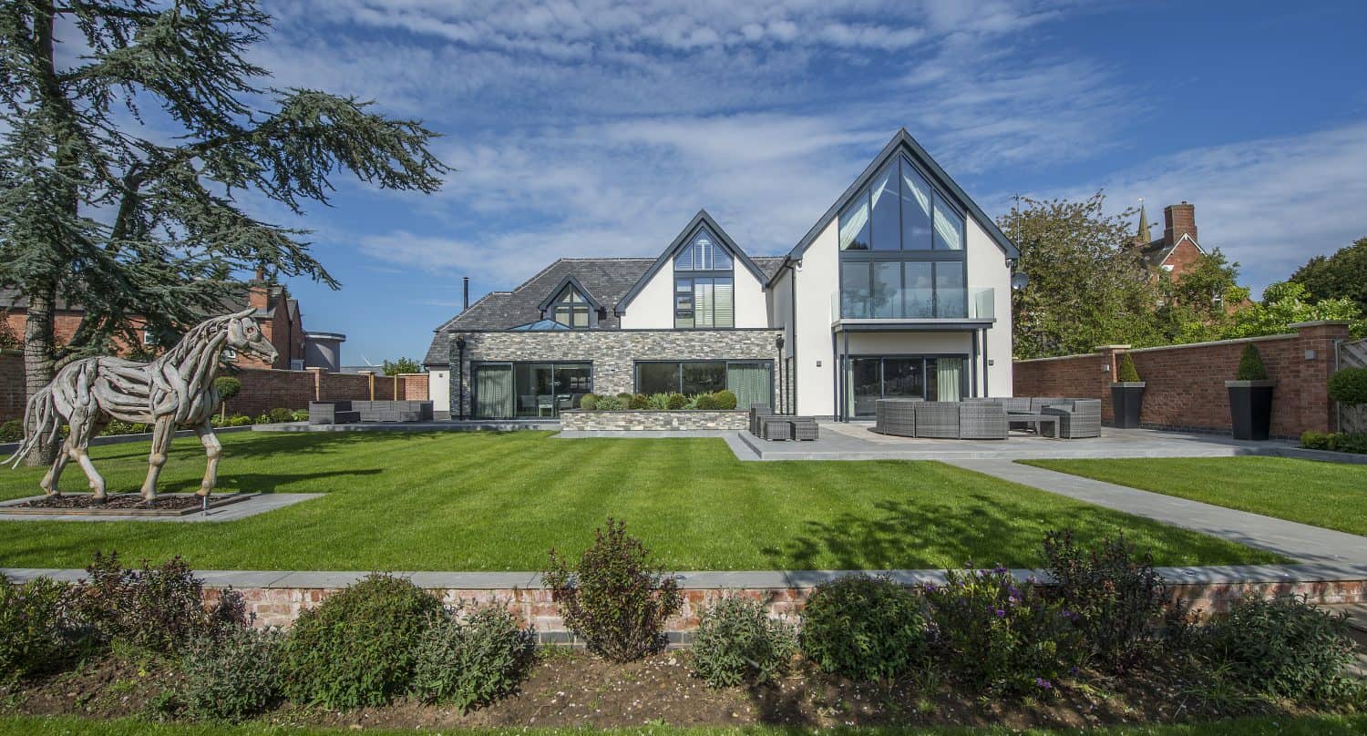 Modern two-story house with large glass windows, sliding doors, and triangular roof lines, set behind a manicured lawn and garden. A metal horse sculpture stands on the left side; the sky is bright with scattered clouds.