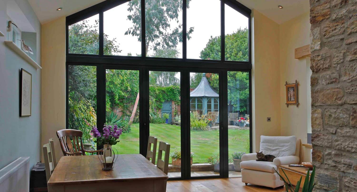 Bright dining and living area with large floor-to-ceiling glass windows overlooking a lush garden with a gazebo; wooden dining table with chairs, a white armchair, and a stone accent wall are visible inside.