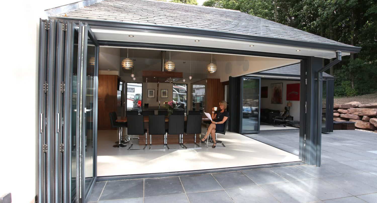 A modern home with large glass folding doors fully opened, revealing a dining area with black chairs, a table, pendant lights, and a woman sitting inside using a laptop. The space opens out to a tiled patio area.