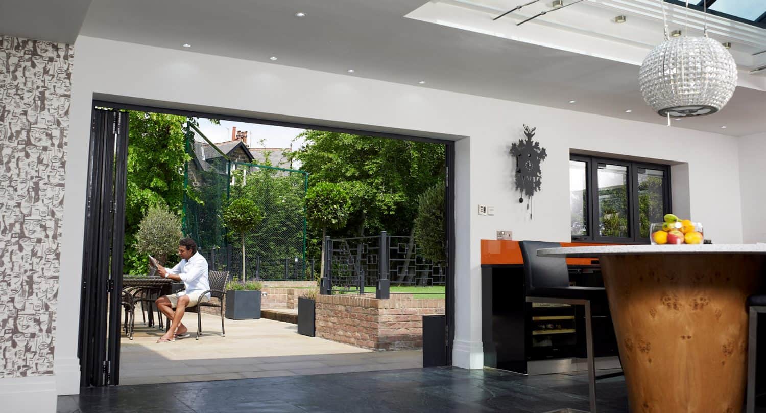 A man sits at a table on a patio outside an open, modern kitchen with a large island, fruit bowl, and chandelier, blending indoor and outdoor living spaces with greenery visible in the garden.