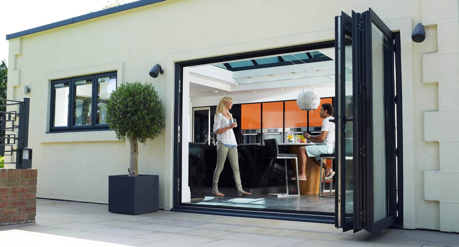 Two women talk in a modern kitchen, one standing inside by a breakfast bar and the other entering from a patio through large open glass doors. The space is bright with a potted tree outside.