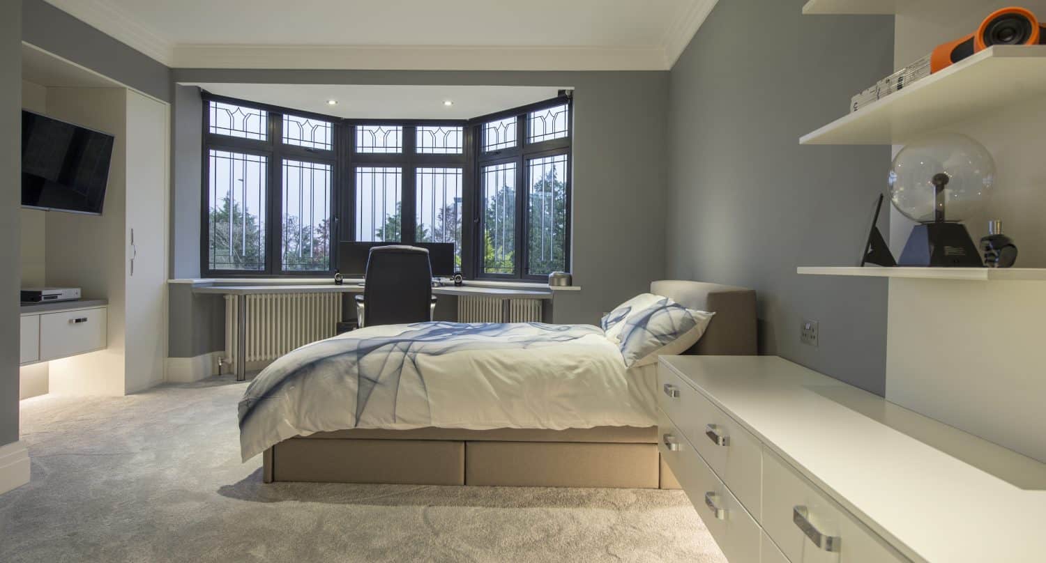 A modern bedroom with a single bed, white furniture, shelves with decor, and a desk by a large bay window featuring bespoke glass solutions that overlook trees. Neutral tones and soft lighting create a calm atmosphere.
