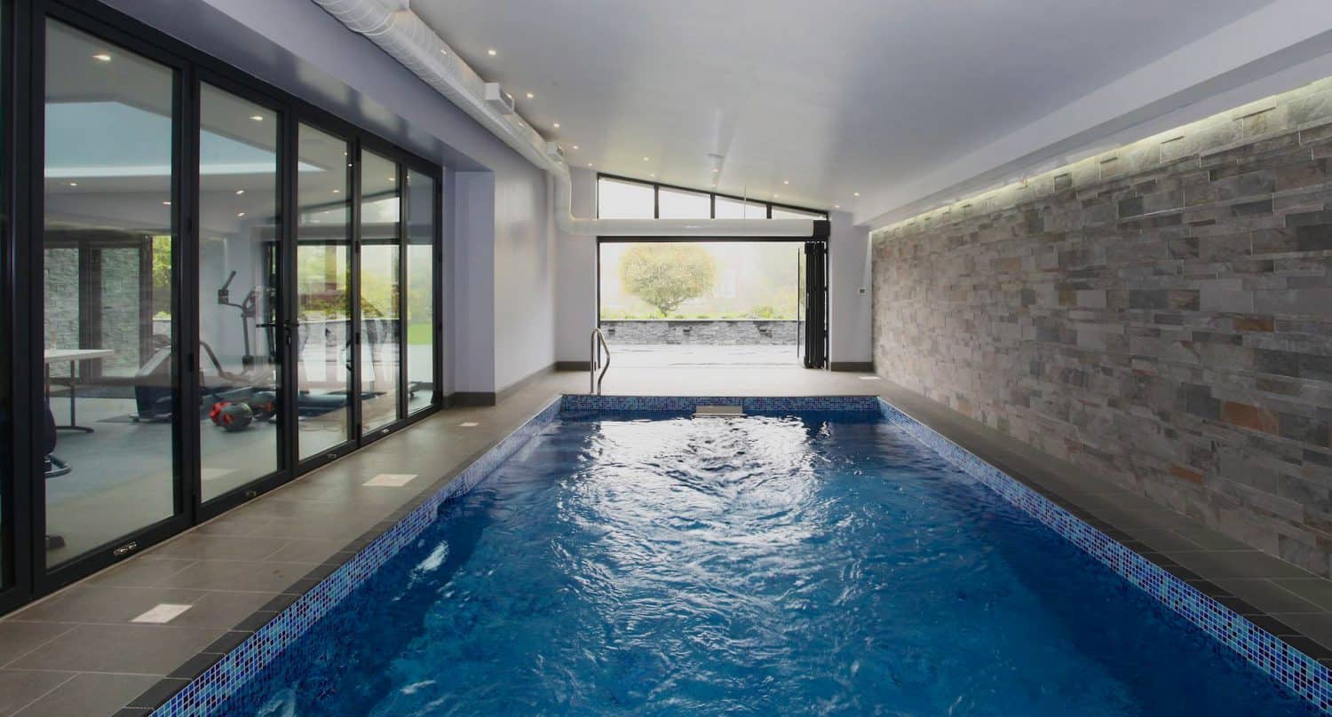 Indoor swimming pool with blue water, bordered by a stone tile wall on one side and floor-to-ceiling glass doors on the other, opening to a gym area and an open patio with greenery outside.