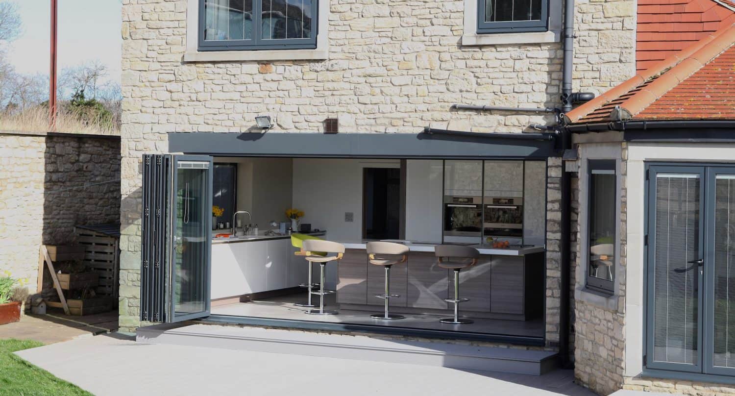 A modern stone house with large bi-fold glass doors fully open, revealing a bright kitchen and four bar stools at a breakfast bar. The outdoor area has a paved patio and some grass visible.