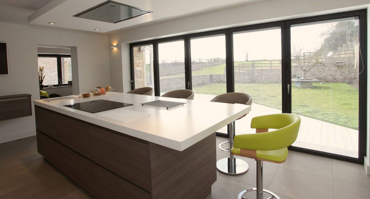Modern kitchen with a large island, built-in stove, three green bar stools, and floor-to-ceiling glass doors opening to a grassy backyard and patio area.