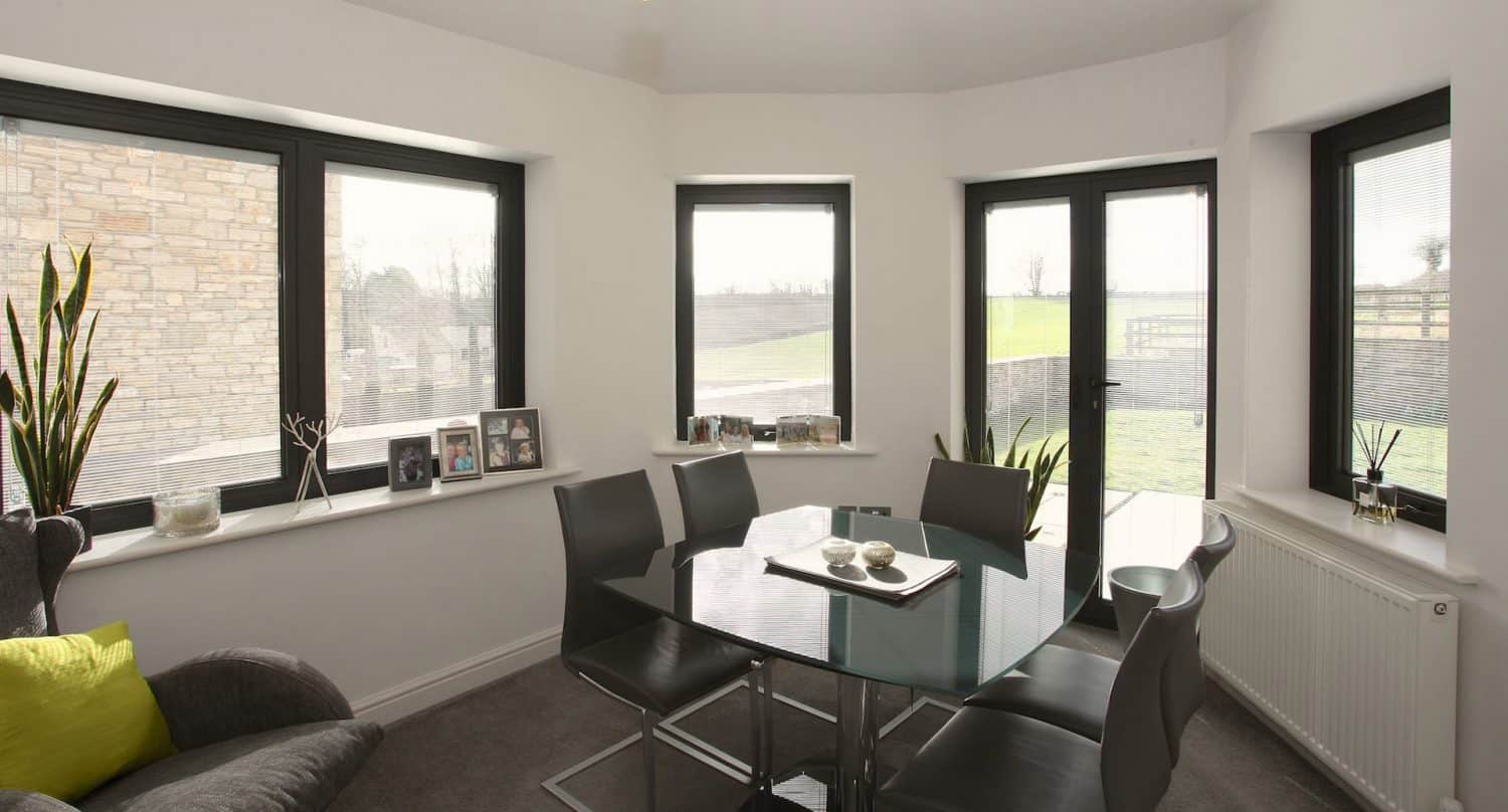 A modern dining area with a glass table, six black chairs, large windows, houseplants, photo frames on the windowsill, and a gray armchair with a green cushion. Daylight streams in through the windows.