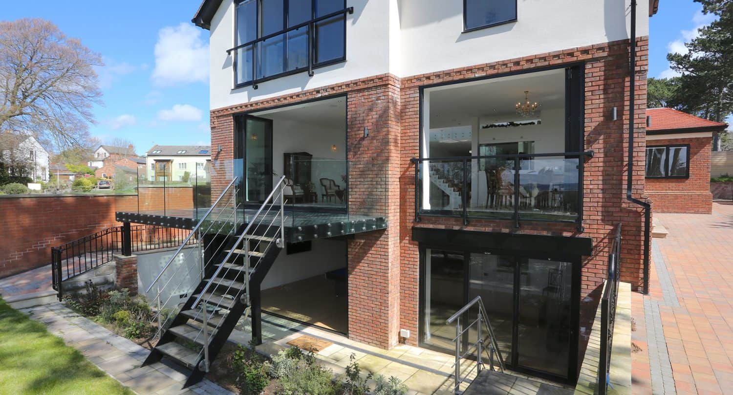 A modern house with large glass windows and brick exterior, featuring metal stairs connecting two outdoor patio areas, with landscaped greenery and a paved driveway visible.
