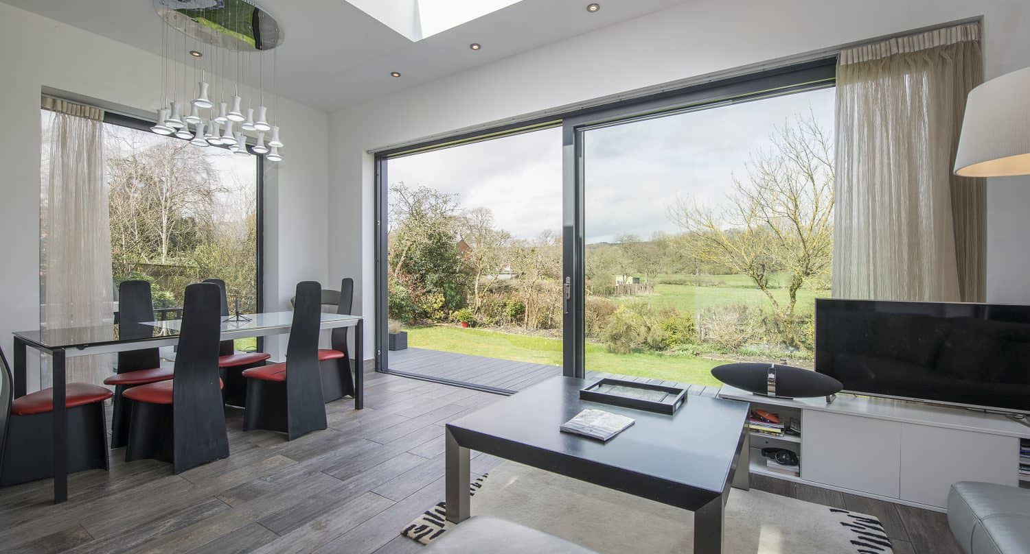 Modern living and dining area with bifold doors overlooking a green garden. Dining table with black chairs, coffee table, TV, and natural light from a skylight and windows create a bright, airy space.