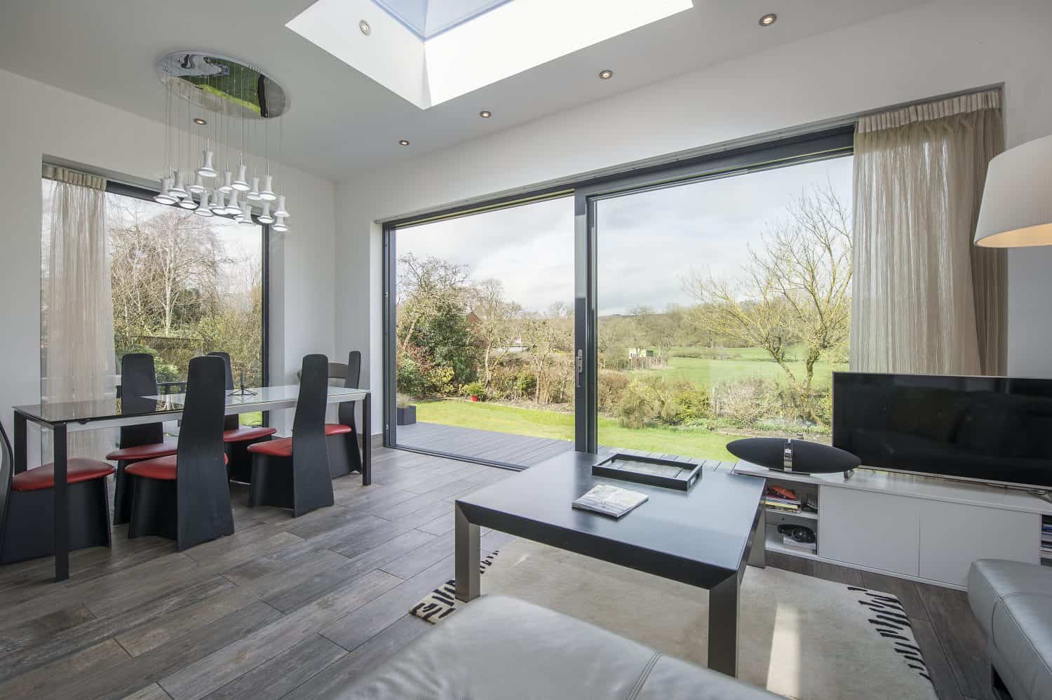 Modern living and dining area with bifold doors overlooking a green garden. Dining table with black chairs, coffee table, TV, and natural light from a skylight and windows create a bright, airy space.
