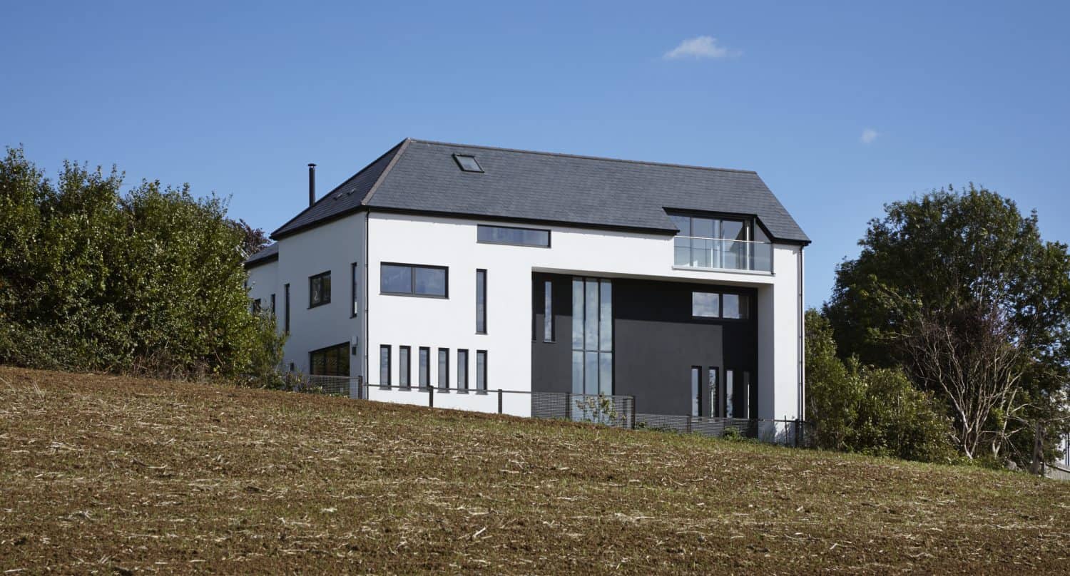 A modern white house with large dark windows and a sloped roof sits on a grassy hill, surrounded by trees, featuring sleek sliding doors under a clear blue sky.
