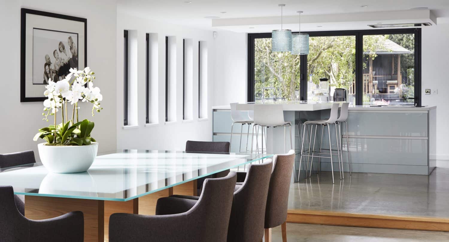 Modern open-plan kitchen and dining area with glass-top table, gray upholstered chairs, bowl of white orchids, bar stools at the island, sliding doors to the garden, large windows, and pendant lights. Bright and airy with garden view.