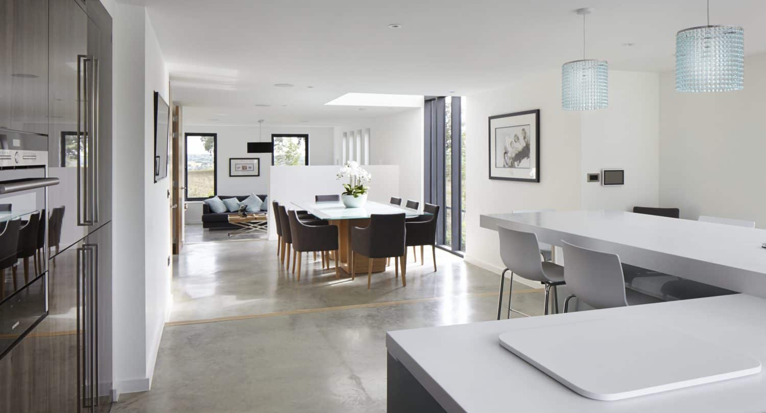 Modern open-plan kitchen and dining area with white countertops, high chairs, hanging blue pendant lights, a large wooden dining table, and a living room visible in the background. Bifold doors allow natural light to beautifully fill the space.