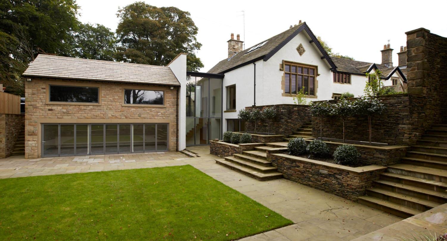 Modern house extension with large glass windows connects to a traditional stone and white house. There are stone steps, a patio, and a neatly trimmed lawn in the landscaped garden.