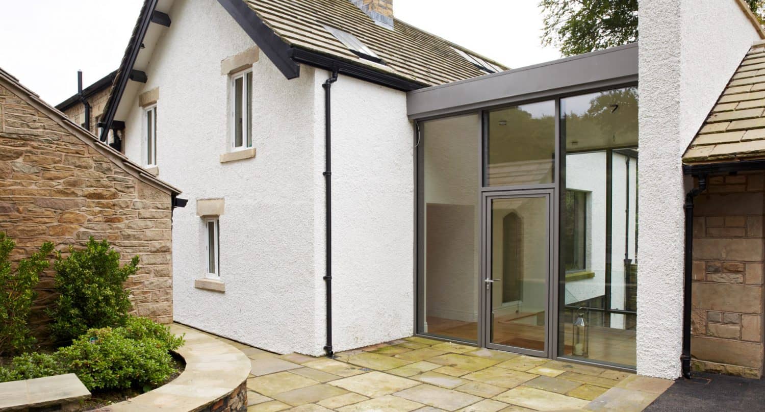 Modern extension with large glass windows and door connects a traditional stone house with a white plastered section, featuring a paved patio and green plants in the foreground.