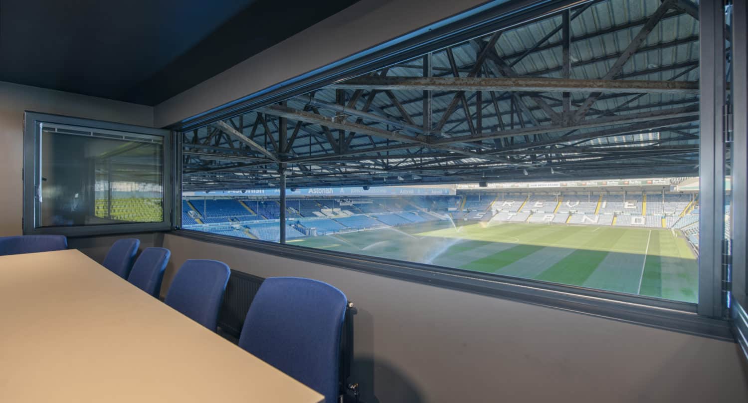 View from inside a private suite with bespoke glass solutions overlooking an empty football stadium, featuring blue seats, green grass, and a partially covered glass roofing structure; a row of chairs and a table are visible in the suite.