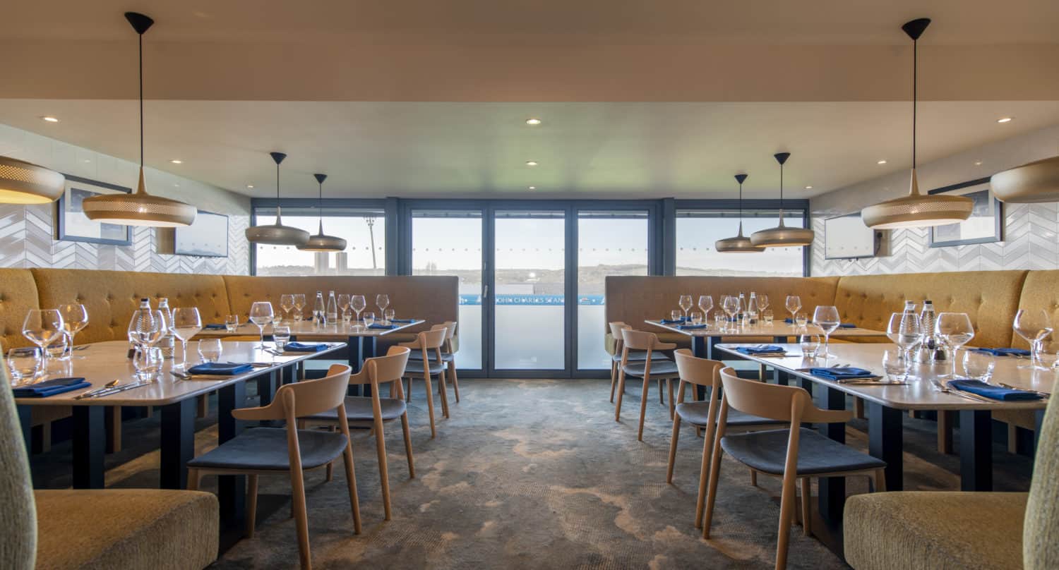 Modern restaurant dining area with wooden tables and chairs, set with glassware and blue napkins. Large windows and sliding doors let in natural light, while pendant lights hang from the ceiling above the tables.