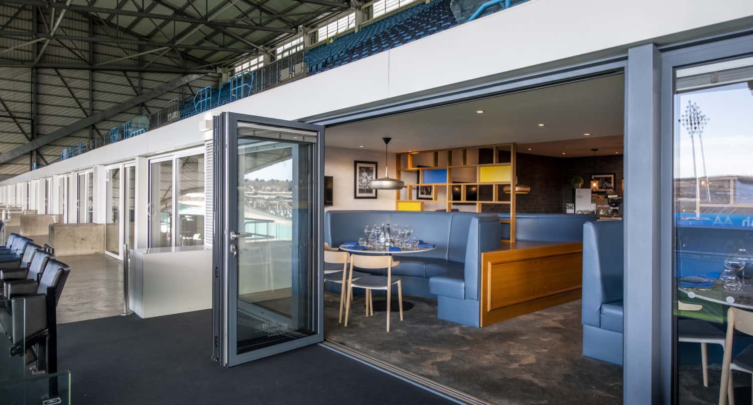 A modern, private stadium suite with blue booth seating, a set dining table, and bespoke glass solutions including large bifold doors open to rows of stadium seats overlooking the field. Bright natural light fills the space.