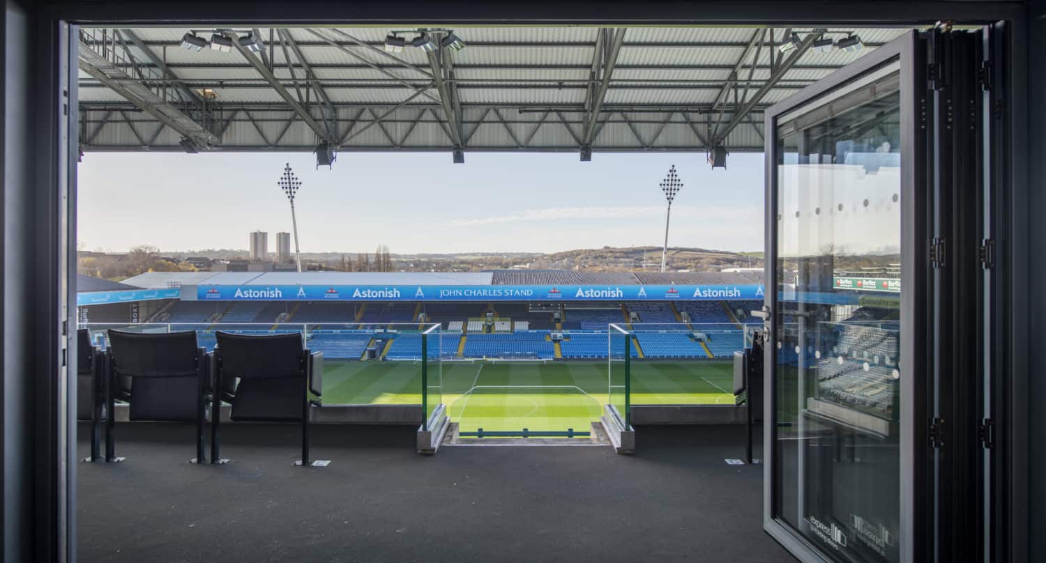 View from a suite overlooking an empty football stadium with blue seats, a well-maintained pitch, and a clear sky visible through large bifold doors.