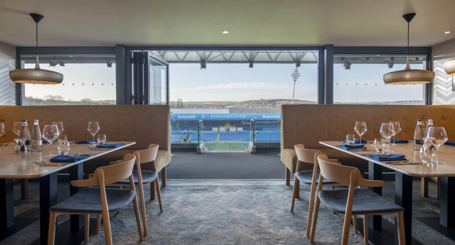 A modern dining area with set tables and wooden chairs overlooks a football stadium through sleek bifold doors, offering a clear view of the field and stands in the distance.
