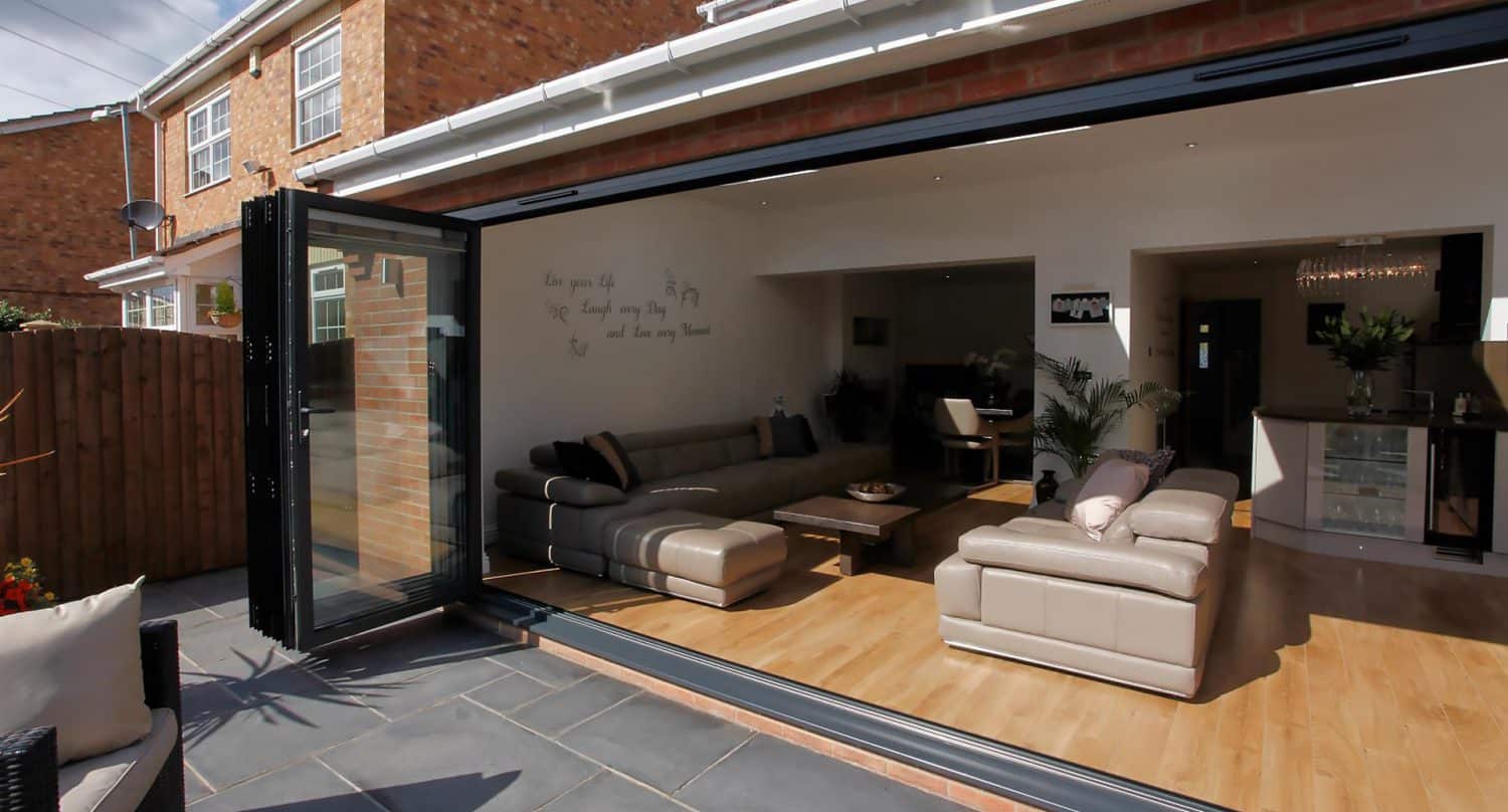 A modern home with large folding glass doors opens to reveal a stylish living room with beige leather sofas, wooden flooring, and contemporary decor connecting to an outdoor patio with gray tiles.