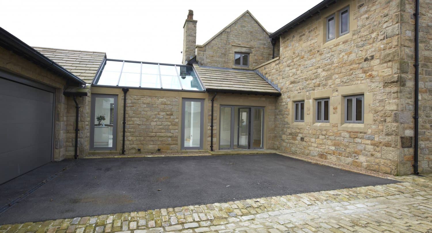 A modern house with stone exterior walls, large windows, a garage on the left, and a paved driveway leading to a glass-covered entrance featuring bespoke glass solutions that seamlessly connect two sections of the building.