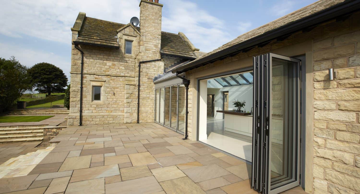 A modern stone house with bifold doors opening onto a spacious stone patio; the bright, open kitchen features skylights and bespoke glass solutions, seamlessly connecting to an outdoor seating area under a blue sky.