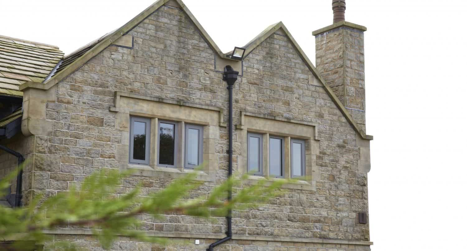 A stone house with two peaked gables, a tall chimney, and multiple rectangular windows. Sliding doors open to the outside, while a leafy green branch is visible in the foreground beneath an overcast sky.