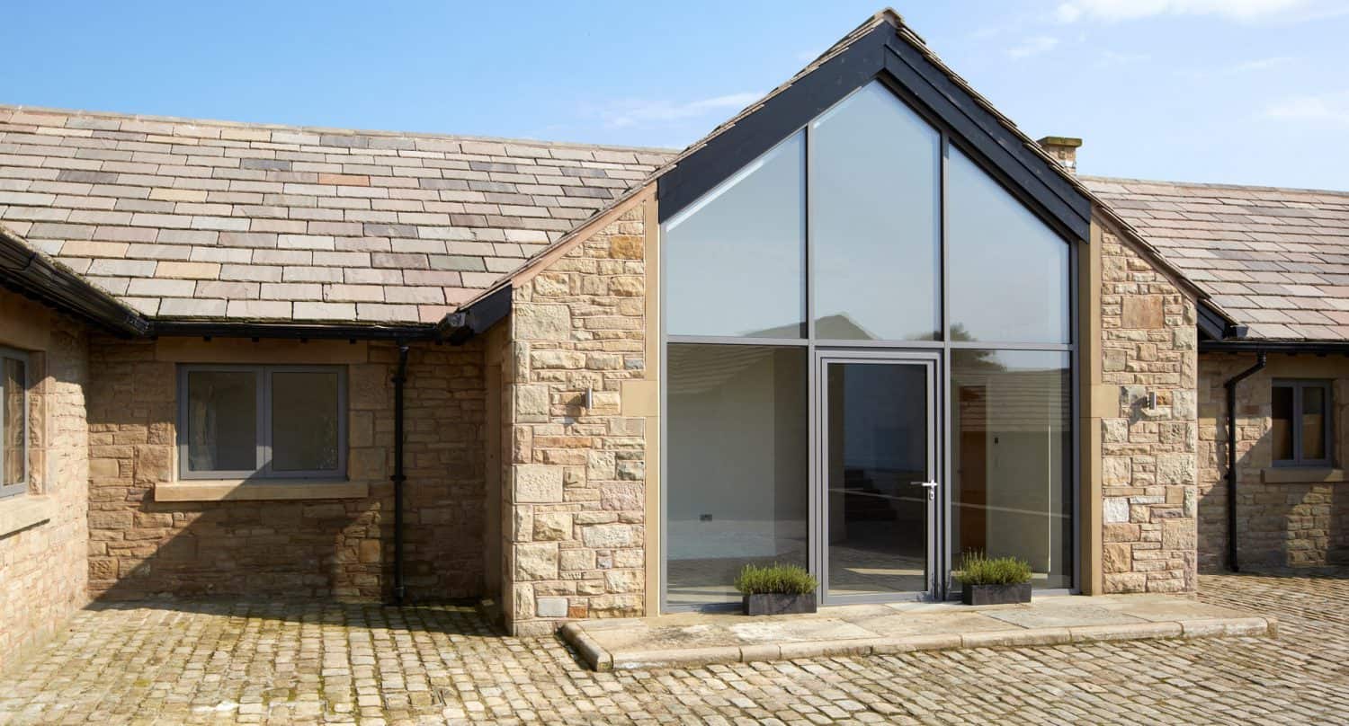 A modern house exterior with stone walls, a high peaked roof, large glass windows, a glass door, and potted plants on a stone patio with a cobblestone driveway.