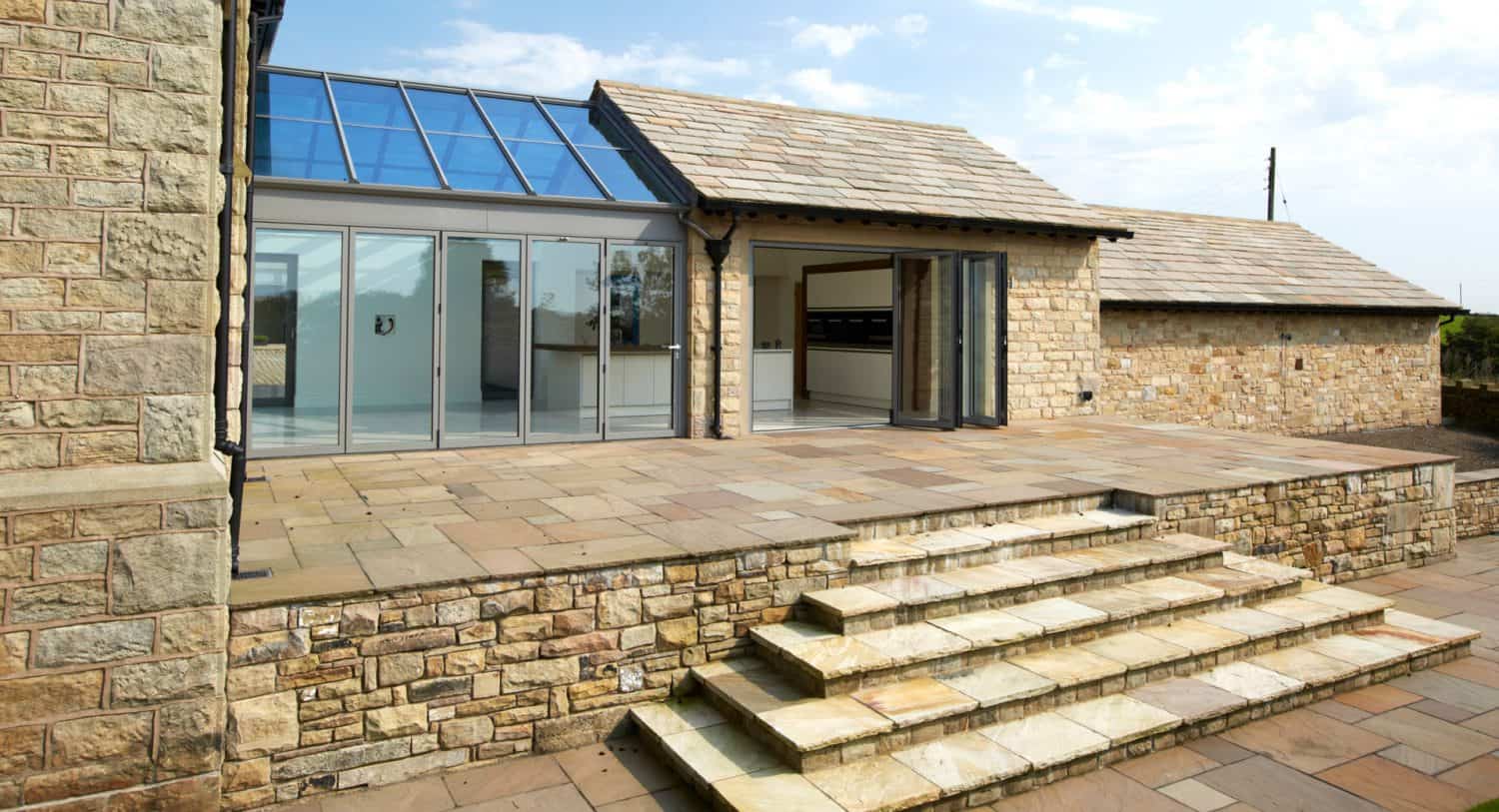 Stone patio with wide steps leading to a modern building featuring large glass doors and a section of sloped glass roof, all surrounded by beige stone walls under a partly cloudy sky.