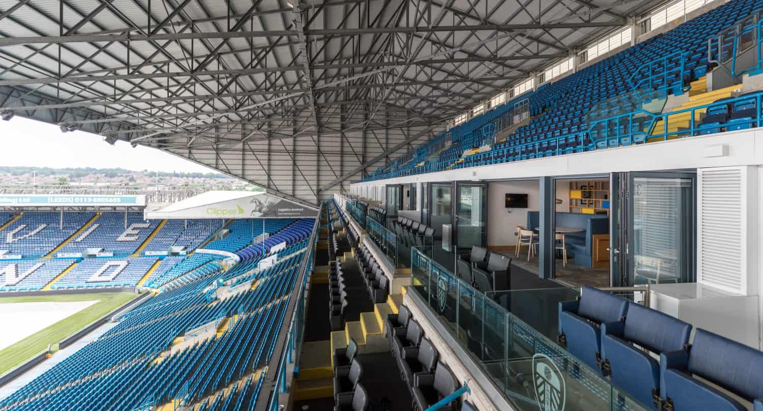 A view of stadium seating and luxury boxes under glass roofing, overlooking the field at Elland Road, home of Leeds United, with empty blue seats and a cityscape visible in the background.