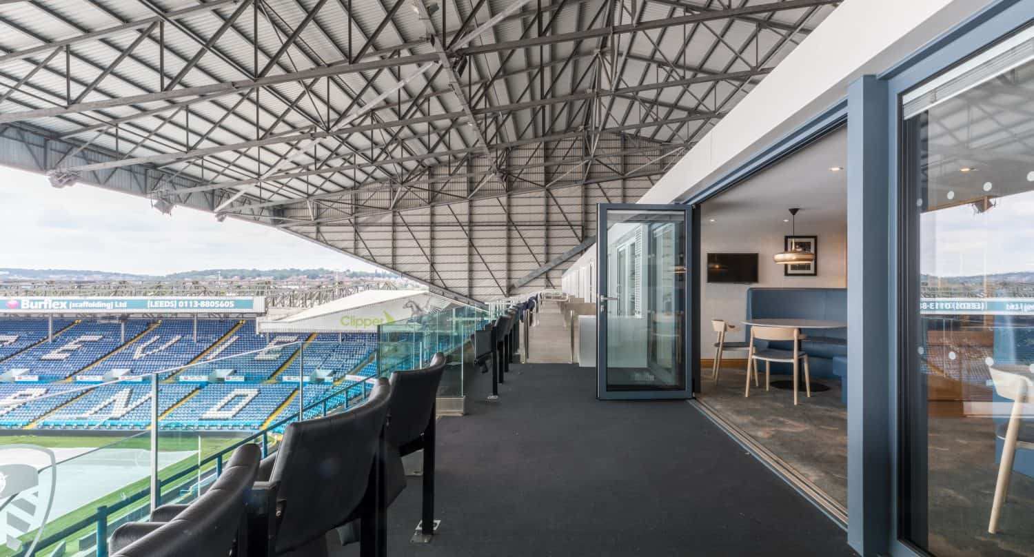 A modern stadium suite features bespoke glass solutions, with sleek glass doors opening onto a private balcony of black seats overlooking the football field. The roof structure and rows of blue seating provide a striking backdrop.