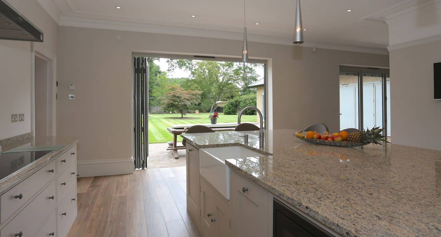 Modern kitchen with granite countertops and fruit bowl, looking out through open folding glass doors to a green lawn where a person is walking near trees. Natural light fills the space.